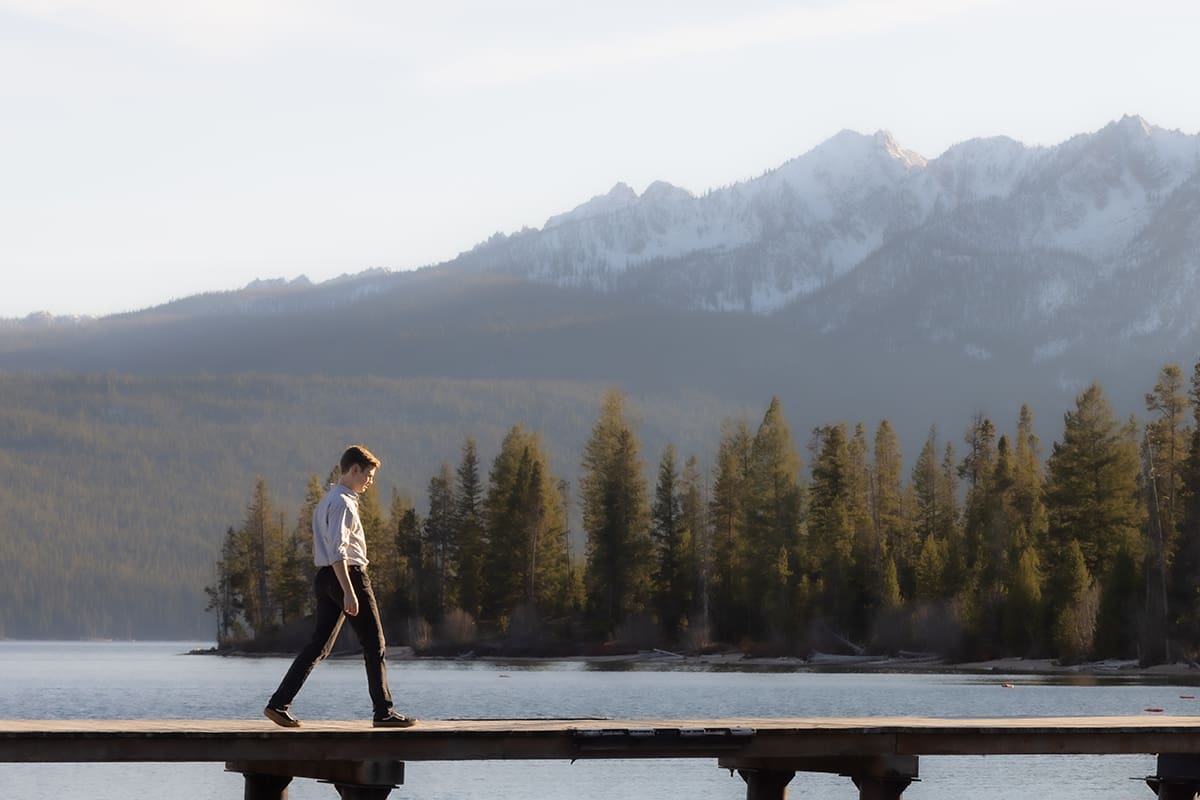 High school senior boy walking down a dock at a lake surrounded by mountains and pine trees.