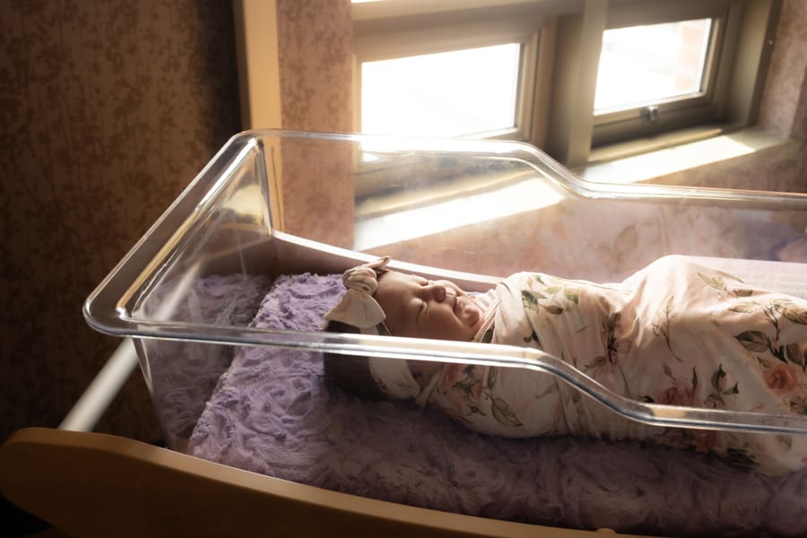 Newborn baby swaddled in a floral blanket with matching headband laying in hospital bassinet.
