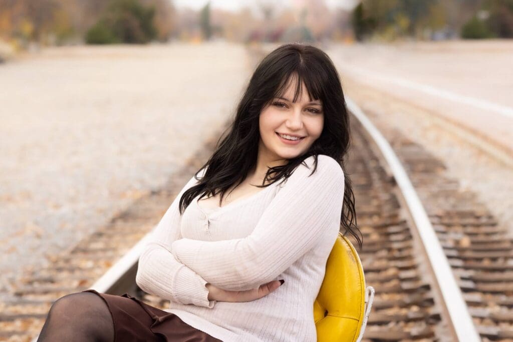 High school senior sitting in a yellow chair placed on railroad tracks for her senior portrait session.