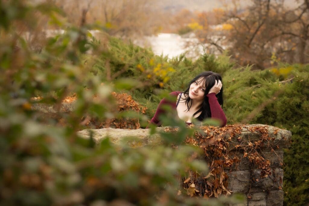 High school senior with her arm resting on an old stone bridge guardrail surrounded by greenery and fall colors.